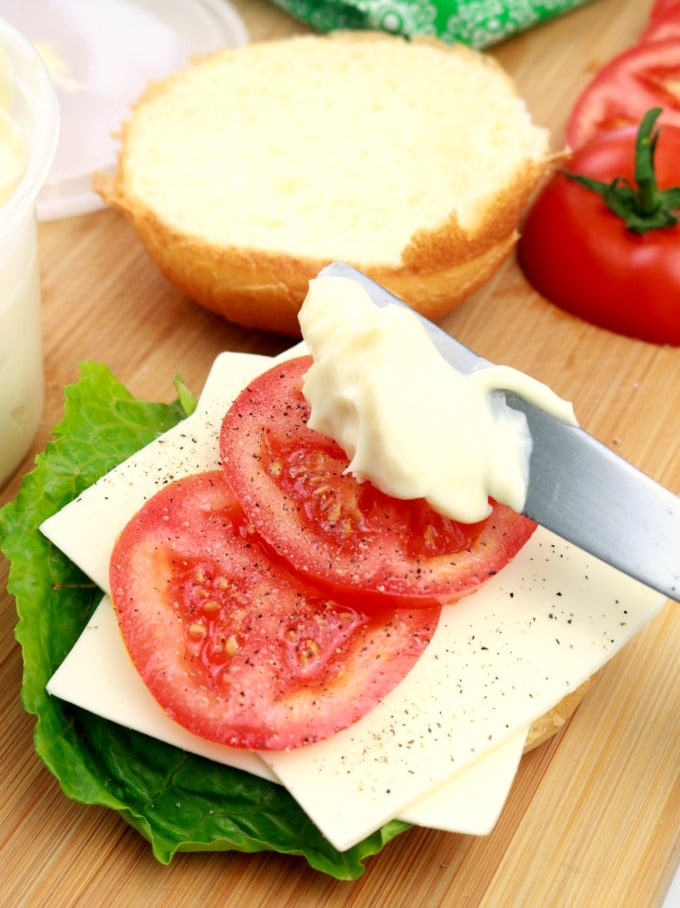 Freshly made mayonnaise being spread over lettuce, tomatoes, and cheese on a brioche bun.