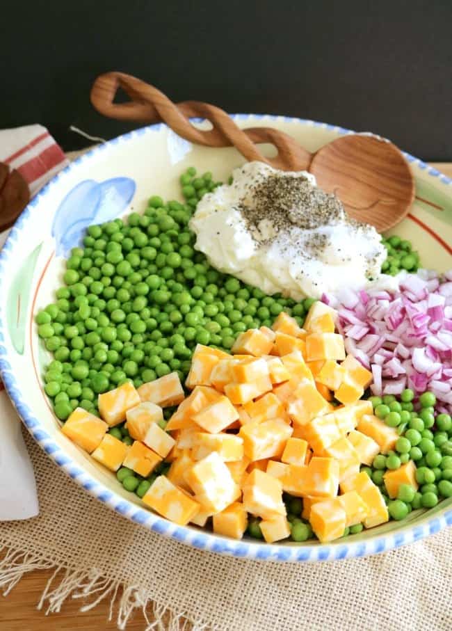 a large serving bowl filled with ingredients for English pea Salad: peas, cheese, red onions, mayonnaise, herbs and seasonings.