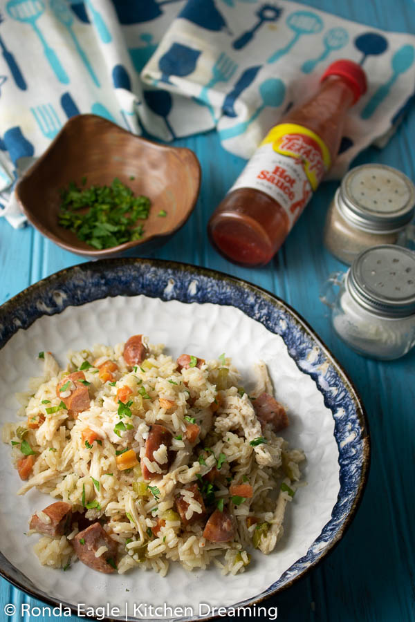 A bowl of southern-style Chicken and Rice on a blue table with a dish of freshly chopped parsley and hot sauce for garnish.