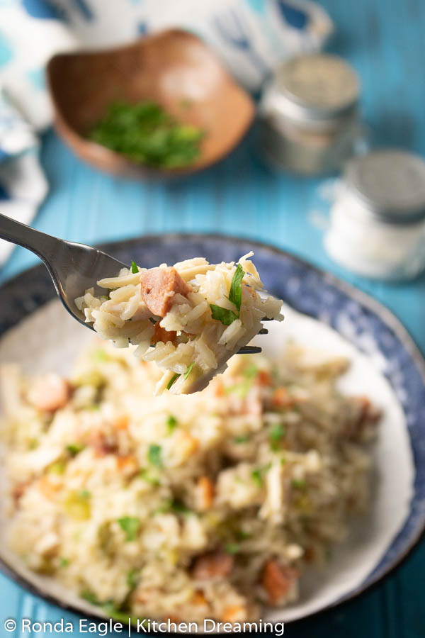 A close-up of a forkful of southern-style Chicken and Rice on a blue table with a dish of freshly chopped parsley and hot sauce for garnish.