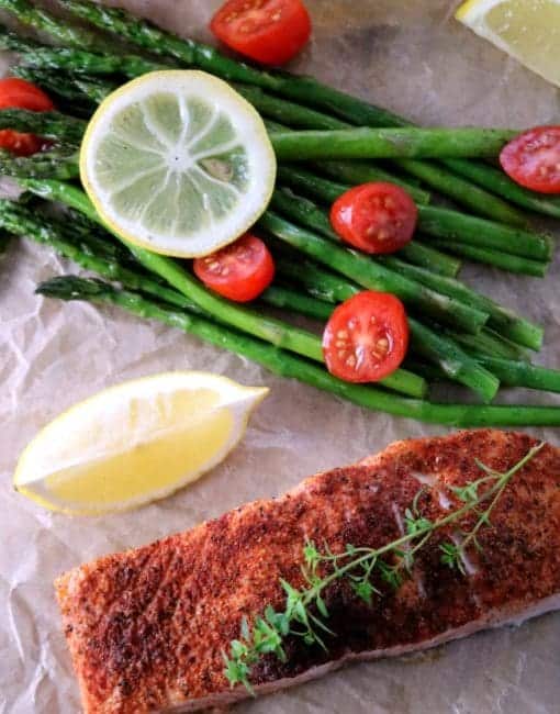 An image of cajun salmon on brown parchment with roasted asparagus, quartered grape tomatoes, and a lemon wedge.