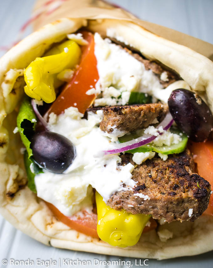 A front on view of some homemade gyro meat wrapped in a fresh pita layered with shredded lettuce, tomatoes, red onion, cucumbers, kalamata olives, pepperoncini peppers, tzatziki sauce, and crumbled feta cheese.