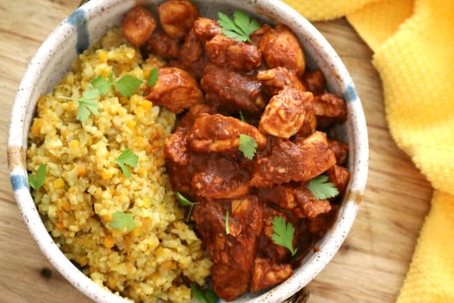 Chicken Vindaloo plated in a clay bowl with cauliflower rice set on a wooden board with a yellow cloth.