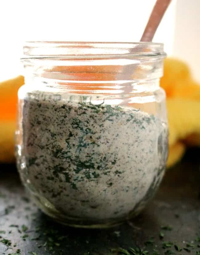 A close-up shot of the side of a glass jar filled with ranch seasoning mix. A yellow towel is in the background. A measuring spoon sticks out of the top of the jar and the jar is sitting on top of a dark slate slab.