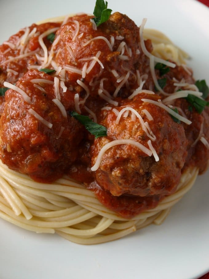 A close up photo of spaghetti and meatballs in marinara sauce on a white plate garnished with Parmesan cheese and torn basil.