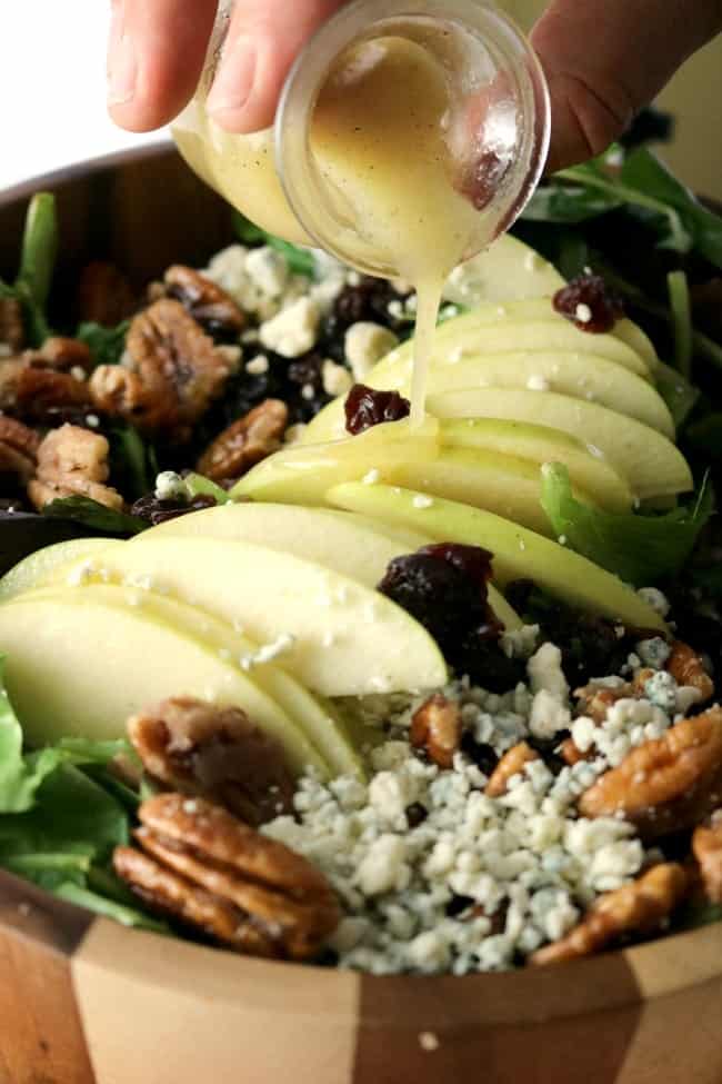 Salad dressing being drizzled over the top of the Apple Pecan Blue Cheese Salad in a wooden bowl.