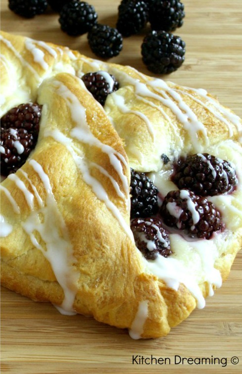 A cheese danish filled with cream cheese and fresh blackberries on a cutting board surrended by more fresh berries.