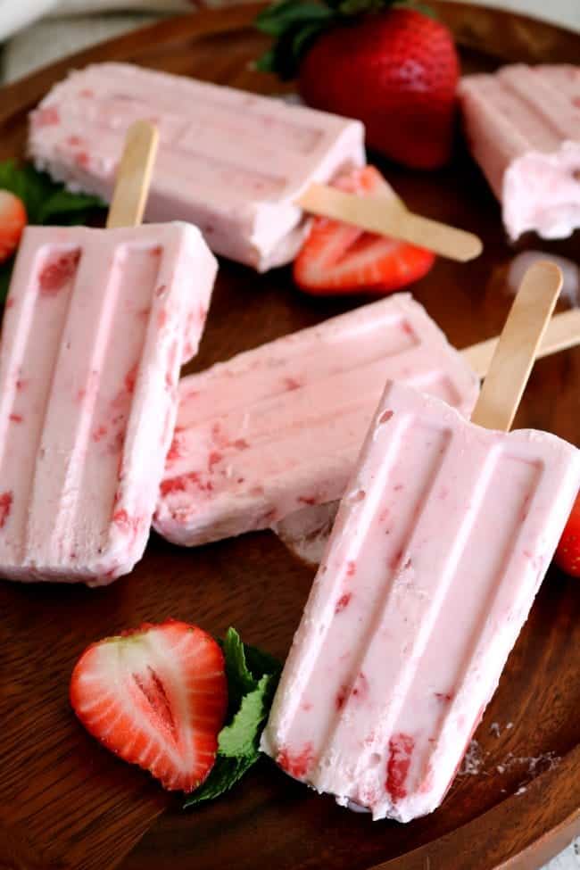 An image of Strawberry cream popsicles on a wooden tray with sliced strawberries. 