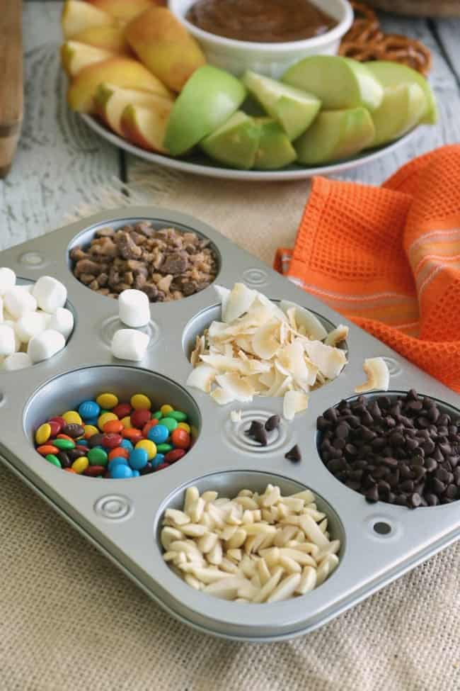 A six-well muffin tin is used to hold various toppings: slivered almonds, mini chocolate chips. toasted coconut flakes, mini marshmallows, and toffee bar crumble.