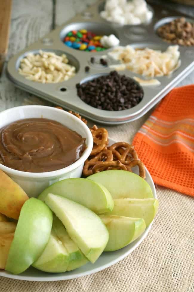 A dish with caramel sauce, sliced apples, and mini pretzels sits in the foreground while a six-well muffin tin holding toppings can be seen in the background.