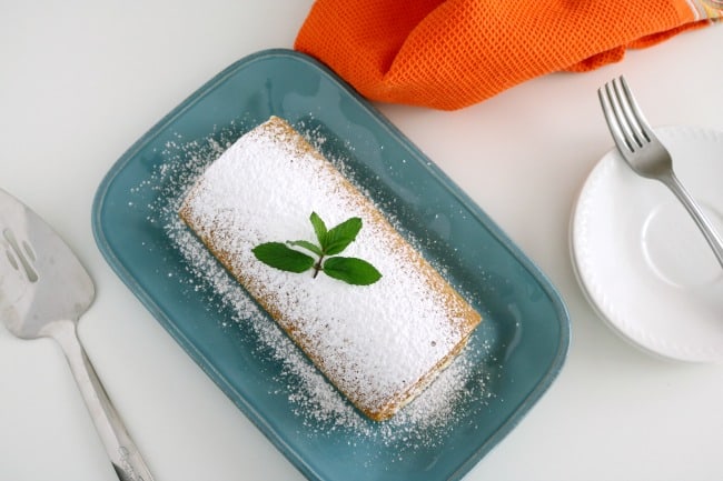 An overhead image of a pumkin cake roll on a blue plate with a cake server with an orange tea towel.