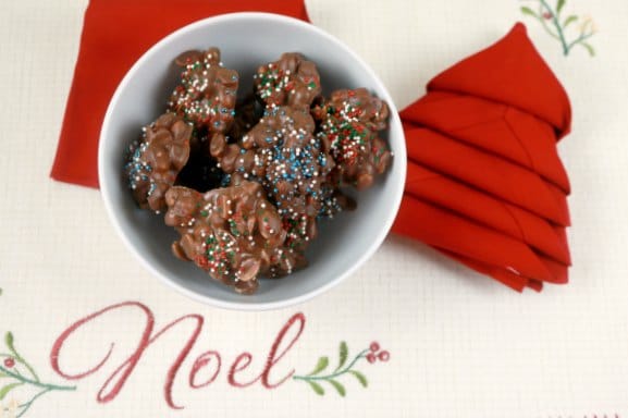 A bowl of slow cooker chocolate peanut clusters on a Noel placemat with a red napkin