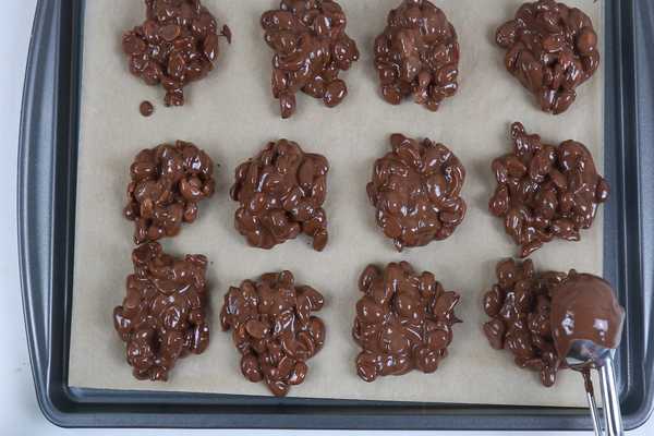 A sheetpan lined with parchent paper that has portioned choclate peanut clusters. The last porting is being added to the bottom right corned of the pan with a small scoop.