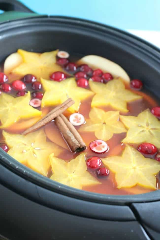 a slow cooker filled with warm holiday punch. slices of apples, oranges, star fruit, cranberries and cinnamon sticks float on top in a spectacular holiday display.