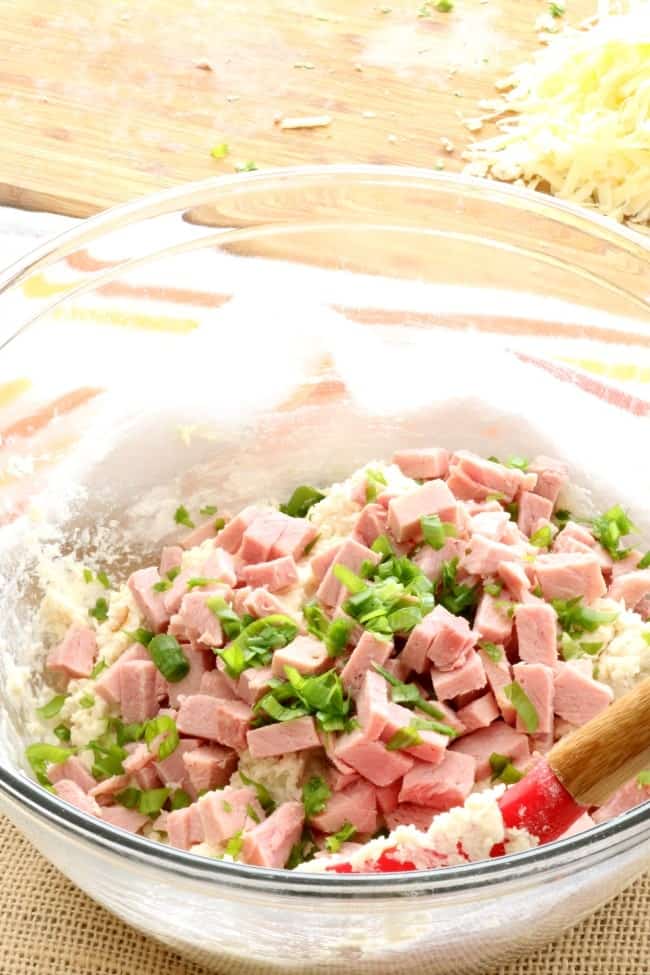 Ham and chives are added into the biscuit batter. Shredded cheese sits in a pile on a cutting board in the background.