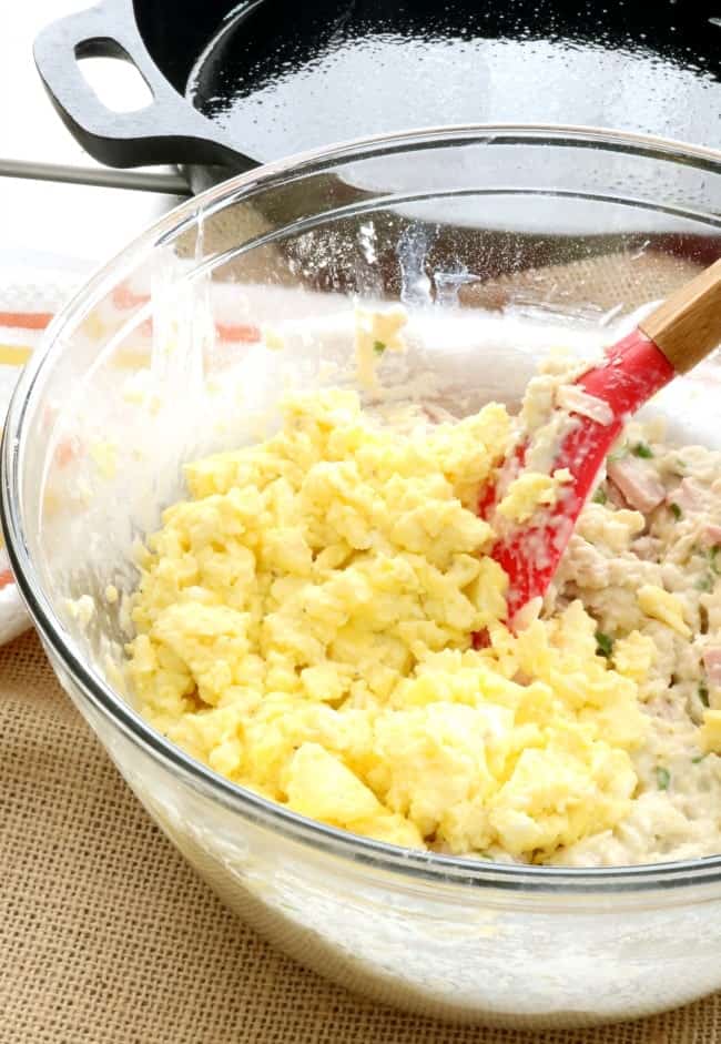 Scrambled eggs are being stirred into a bowl of ham and cheese biscuits. a prepared cast iron skillet awaits in the background.