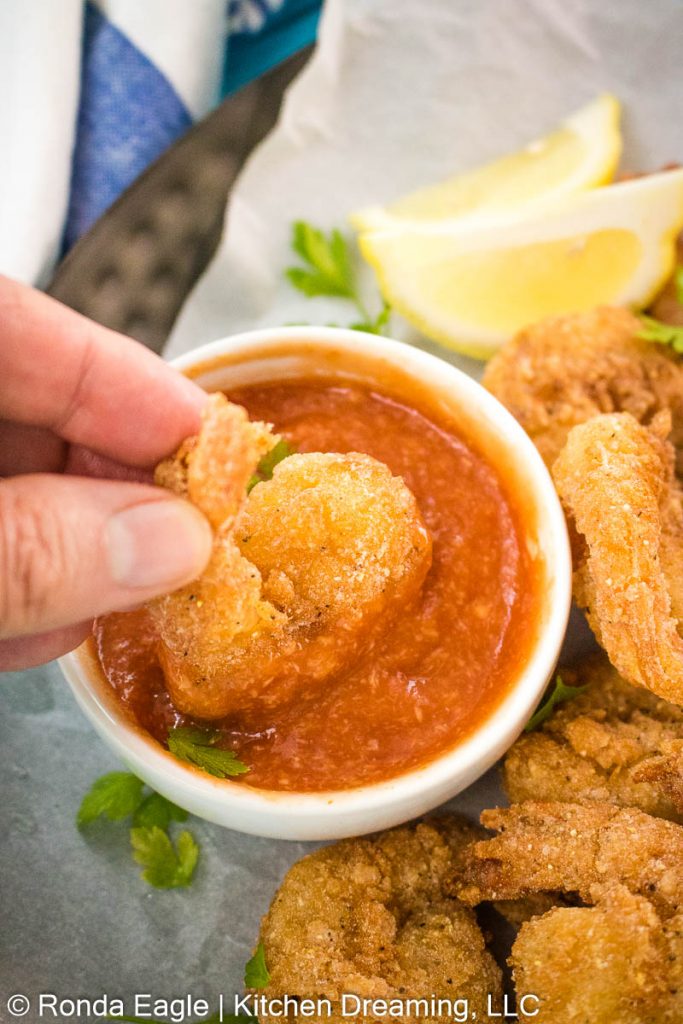 A piece of fried shrimp is being dipped into a small bowl of homemade cocktail sauce.