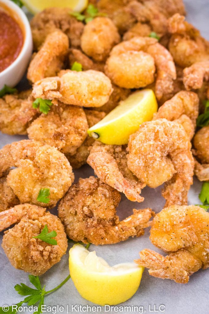 An up-close image of a basket of fried shrimp with a dish of homemade cocktail sauce for dipping. Lemon wedges and a sprinkling of parsley are on the tray as garnishes.