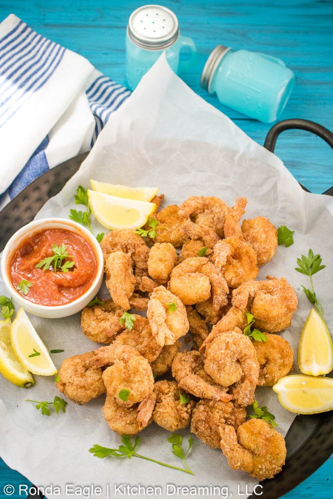 A basket of fried shrimp with a dish of homemade cocktail sauce for dipping. Lemon wedges and a sprinkling of parsley are on the tray as garnishes.