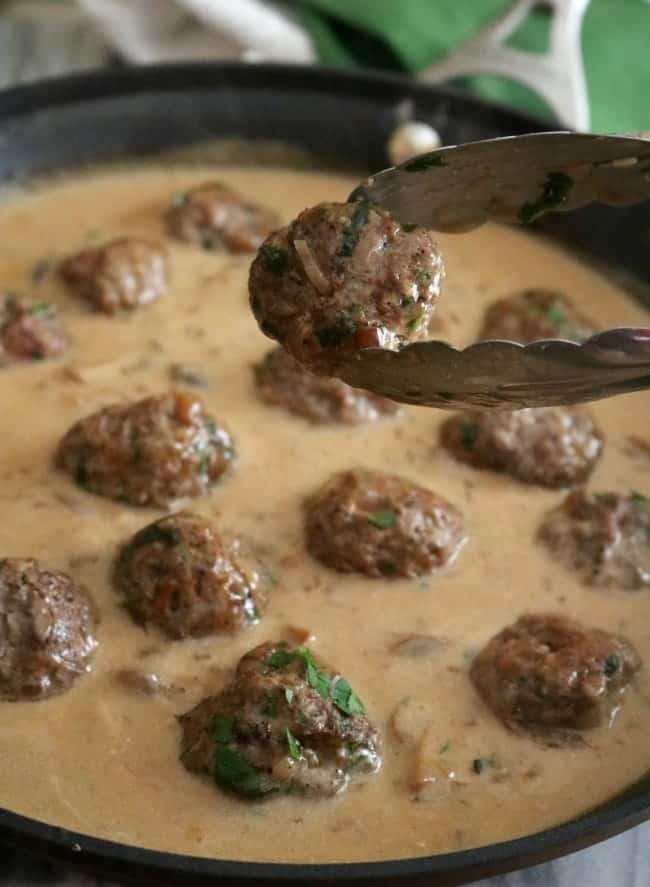 A pair of tongs holds up a meatball over the pan of luscious, creamy, brown gravy.
