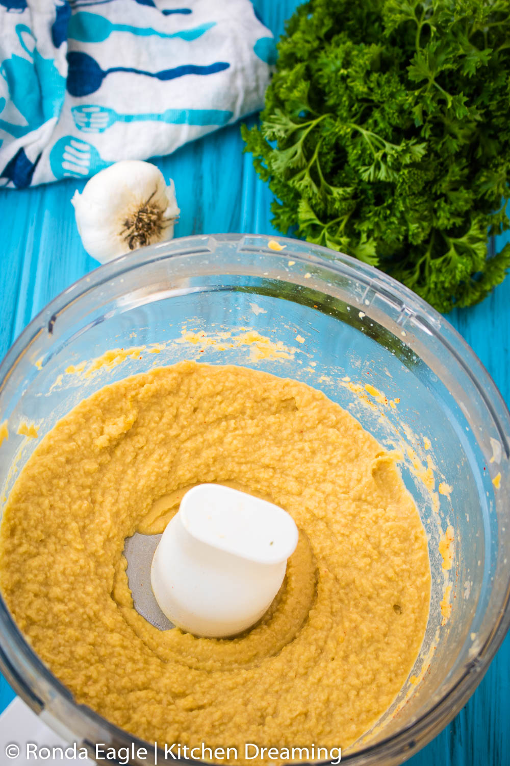 An image of chicken pea hummus that has been blended. It is in the bowl of the food processor. Curly parsley, a head of garlic, and a blue and white patterned towel are set in the background. 