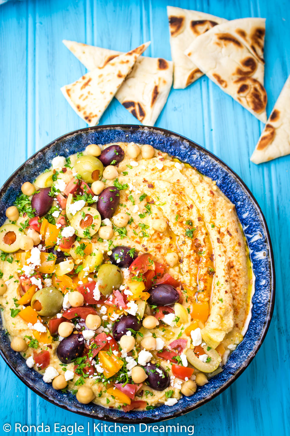 An image of an overhead shot of a bowl of loaded hummus. What makes this hummus different is that it is prepared without tahini paste. The top of the bowl has been garnished with chopped grape yellow and red grape tomatoes, Kalamata olives, green olives, additional chick peas and a sprinkling of crumbled feta and minced parsley. It was then drizzled with olive out and a dusting of paprika and red pepper flakes. 