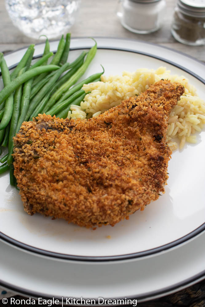 A table setting with an air fried pork chop, steamed green beans, and rice on a white plate.