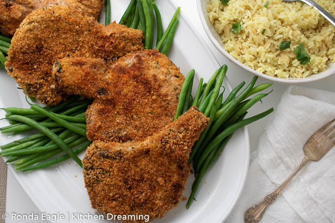 A horizontal photo of a plate of crispy breaded pork chops that have been cooked in the air fryer. They are surrounded by freshly steamed green beans. 