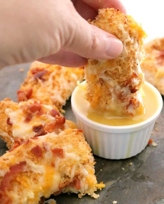 a hand dipping a cheddar bacon topped chicken tender into a white ramekin of homemade honey mustard on a dark slate tile.
