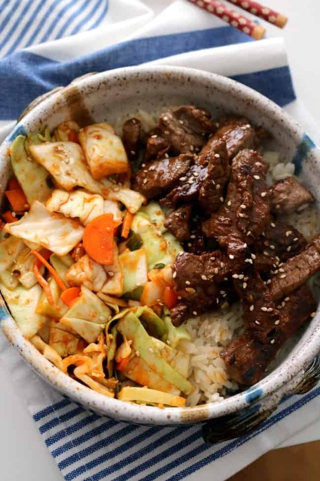 Korean Beef Bowls with kimchi over rice sitting on a blue and white cloth with chopsticks