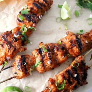 Korean chicken skewers that have been grilled - they are laying on a brown paper with wedges of lime and slices scallions.