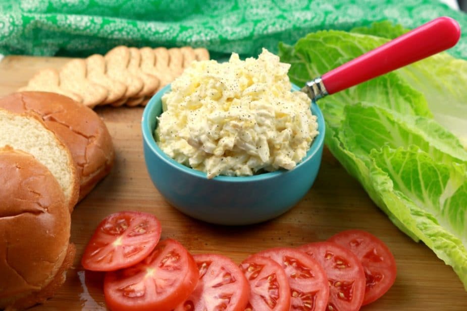 a horizontal image of egg salad on a wooden board. the bowl is surrounded by lettuce, tomatoes, brioche rolls, and crackers