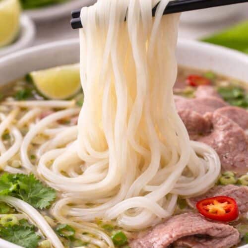 Fresh homemade rice noodles lifted with chopsticks over a bowl of soup, showing smooth gluten-free noodle texture.