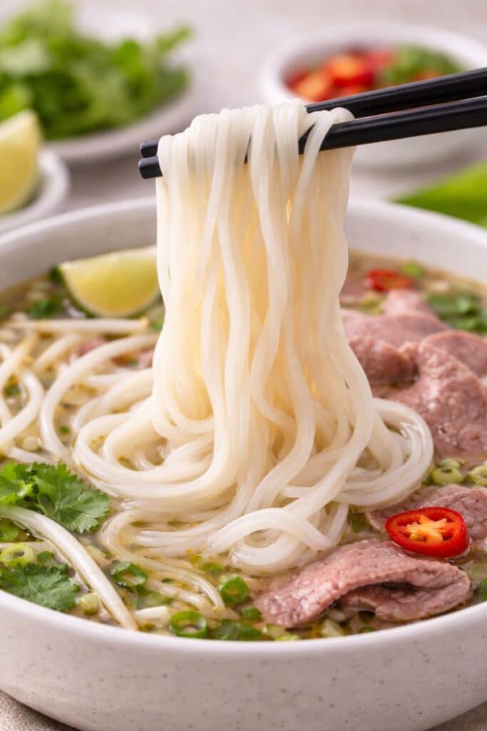 Fresh homemade rice noodles lifted with chopsticks over a bowl of soup, showing smooth gluten-free noodle texture.