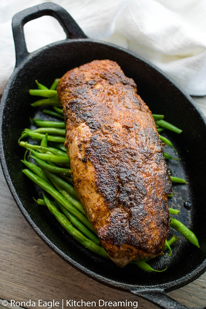 An oblong cast iron skillet filled with roasted green beans and a whole pork tenderloin.