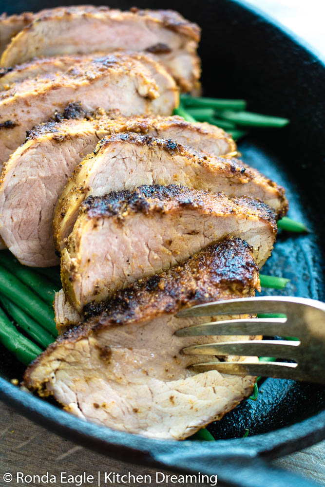 A close up image of an oblong cast iron skillet filled with roasted green beans and sliced pork tenderloin. A serving fork pierces the foremost piece of meat and juices are running out of the meat.