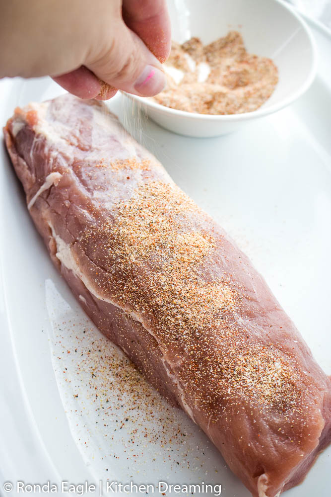 A raw pork tenderloin on a white plate being seasoned with dry rub.