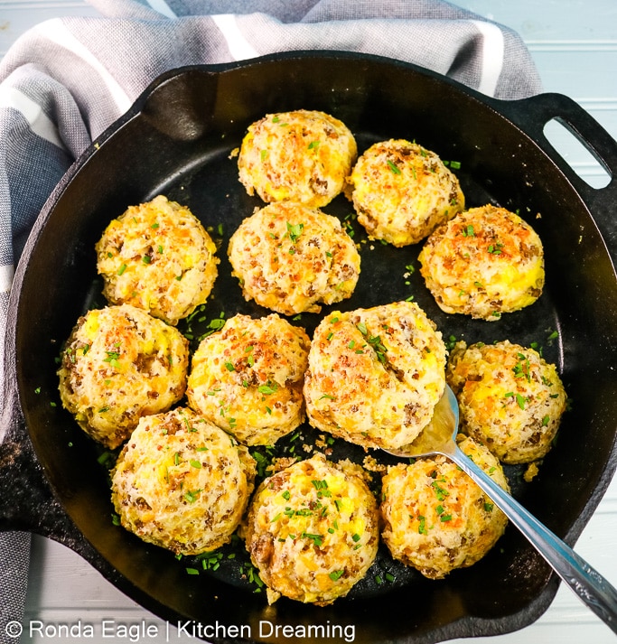 A pan of sausage biscuits that has two biscuits missing. A silver serving spatula rests under a biscuit and on the edge of the cast iron pan.