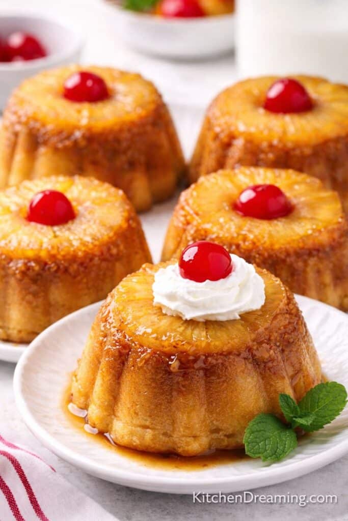 Individual pineapple upside-down cakes in mini bundt shapes with caramelized pineapple rings and cherries, served on plates.
