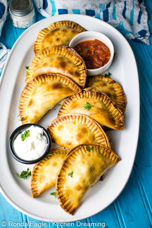 A plate of baked beef empanadas with sour cream and salsa for dipping.