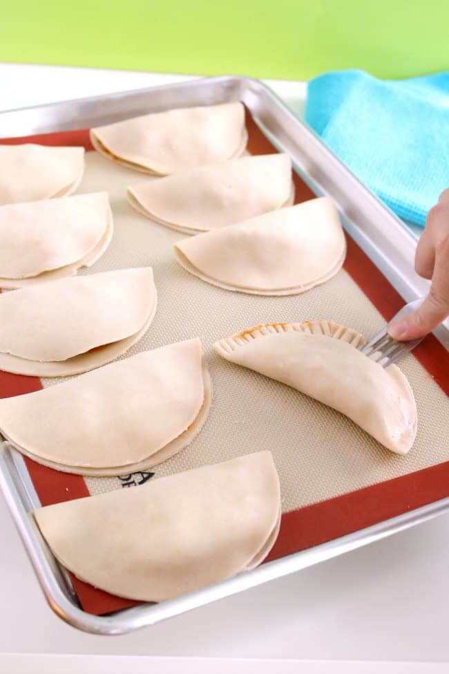 A tray of empanadas which have been filled and the edges are being crimped with a fork. 