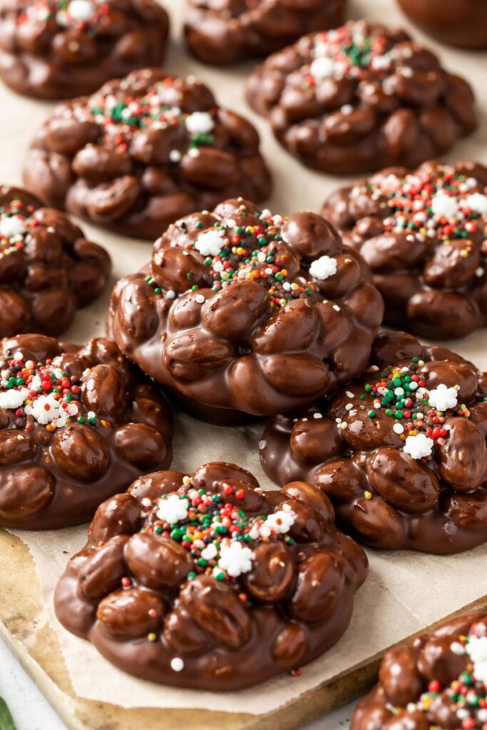 Crockpot chocolate peanut clusters on parchment paper, coated in glossy chocolate and topped with holiday sprinkles. Easy slow cooker candy that sets up firm, sweet-and-salty peanut chocolate clusters.