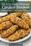 Crispy air fryer garlic parmesan chicken tenders stacked on a white plate with parsley, extra Parmesan, dipping sauces, and lemon wedges.