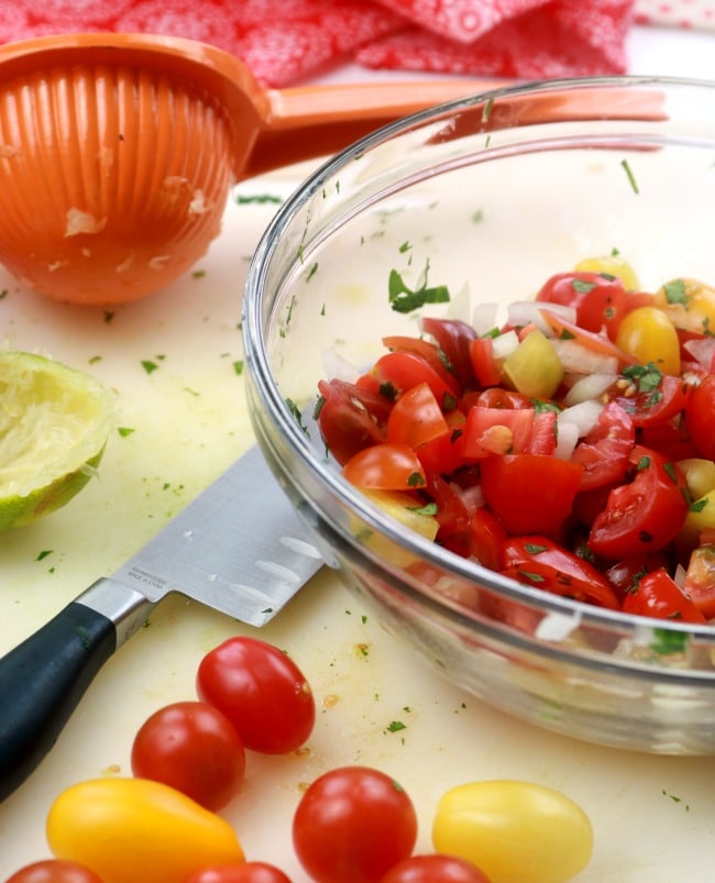 A bowl of fresh pico de gallo on a cutting board with a lime, a citrus juicer, and some fresh grape tomatoes.