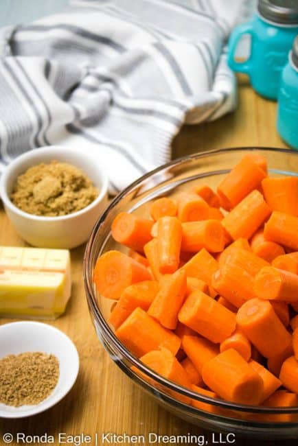 A medium-sized glass mixing bowl filled with peeled and cut carrots. Next to the bowl are some ingredients to make glazed carrots: brown sugar, butter, ginger, and fresh nutmeg.