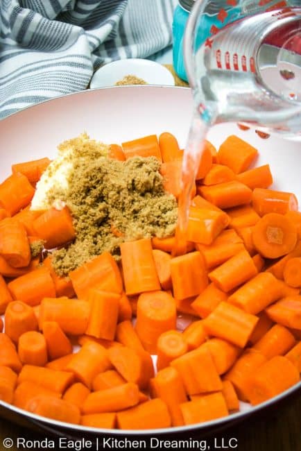 A pour shot of water being added to the skillet of carrots. In the background of the pan, you can see the butter and brown sugar on top of the carrots.