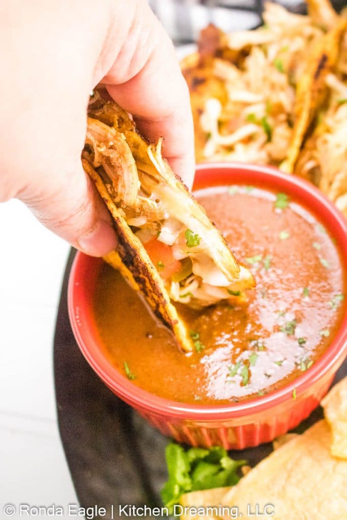 A hand is dipping a birria taco into a dish of adobo consomé.