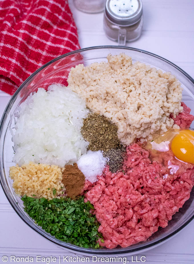 A glass bowl containing the ingredients for  Greek Meatballs; beef, onions, garlic, parsley, egg, herbs & spices. For a full list, visit the recipe card.