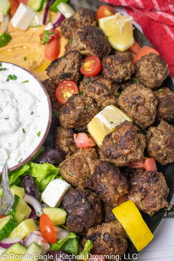 A metal platter filled with a Greek-inspired feast of  Greek Meatballs [also called Keftedakia or Keftedes], Tzatziki Sauce, Hummus, Greek Salad, and and soft, warm fresh pita slices. 