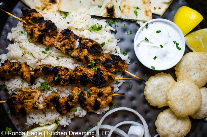 A black hammered metal serving tray filled with Grilled CHicken skewers on a bed of rice, sweet onions, lemon wedges, and raita.